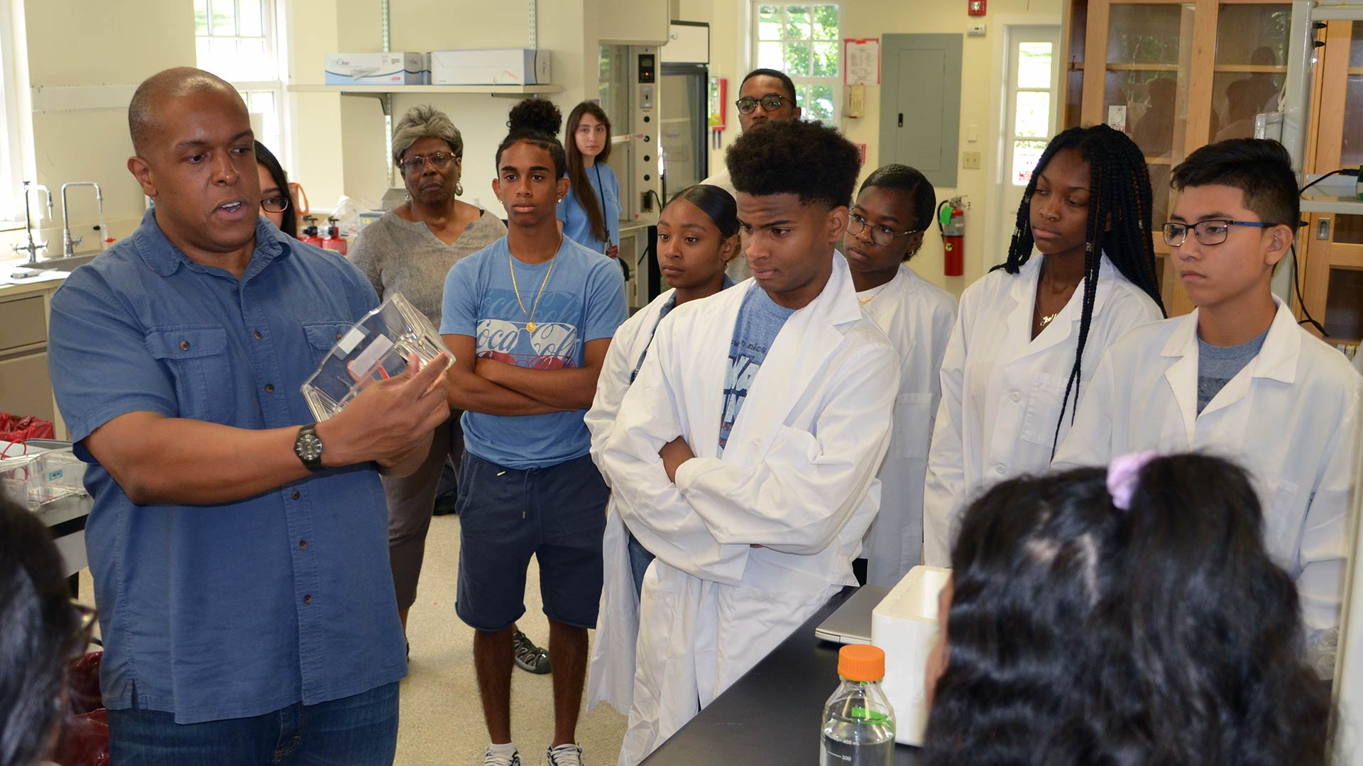 Assistant Director Jason Williams demonstrates a scientific object to attentive students in a lab, some wearing lab coats.
