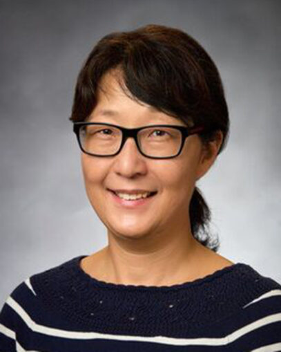 A photograph of Seung Yon (Sue) Rhee smiling. She is wearing glasses with black frames, and a black sweater with white horizontal stripes.