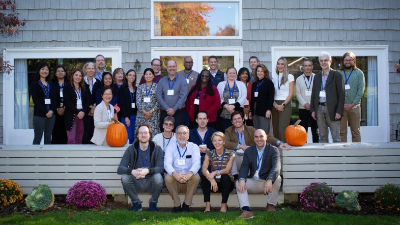 31 people positioned in four rows on the wood back patio of the Banbury Center's conference room. All meeting participants are wearing blue lanyards with name tags and are smiling.