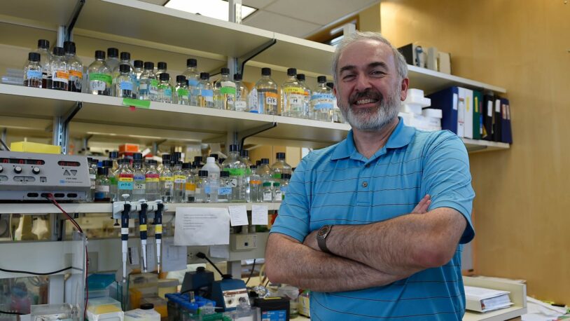 A smiling man in a blue shirt stands with arms crossed in a science lab filled with bottles and equipment.