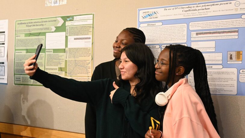 Three high school students stand together smiling as they take a selfie in front of DNALC scientific research posters displayed on a wall behind them. One student gives a thumbs-up gesture while another wears headphones around their neck.