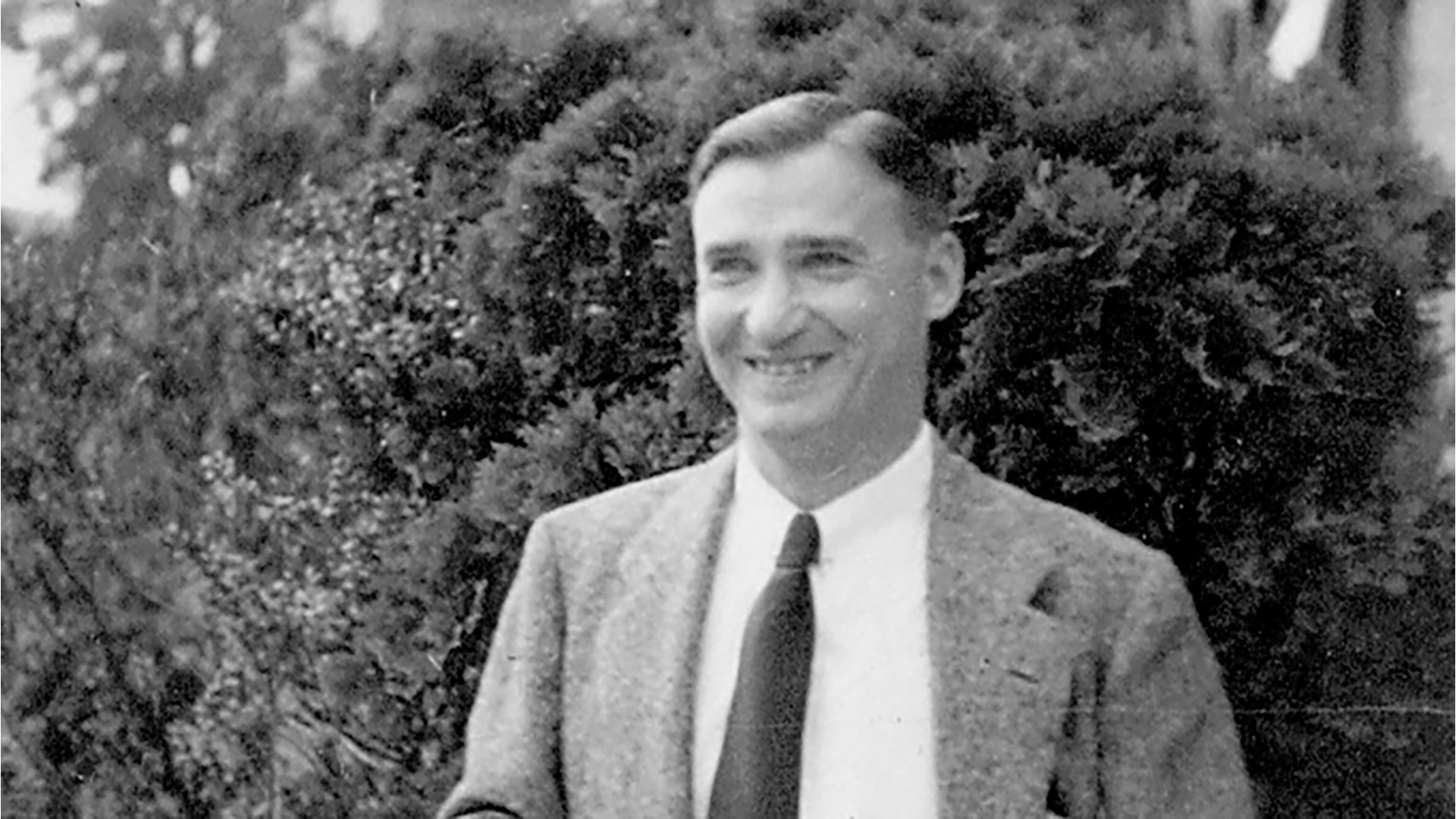 A man in a suit and tie smiles outdoors in front of leafy shrubs, in a black-and-white photo.