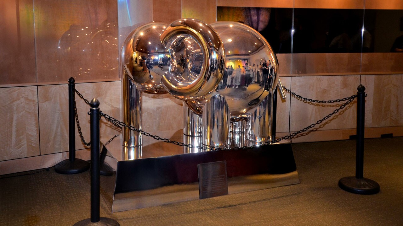 Shiny metallic elephant sculpture displayed indoors, surrounded by black stanchions and chains.