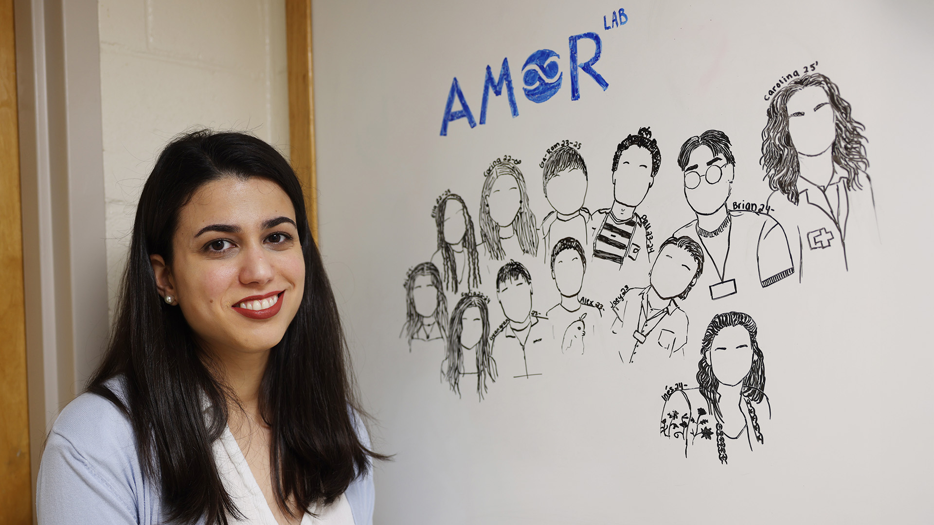 Amor Vegas smiling next to a whiteboard with AMOR LAB and hand-drawn sketches of people underneath.