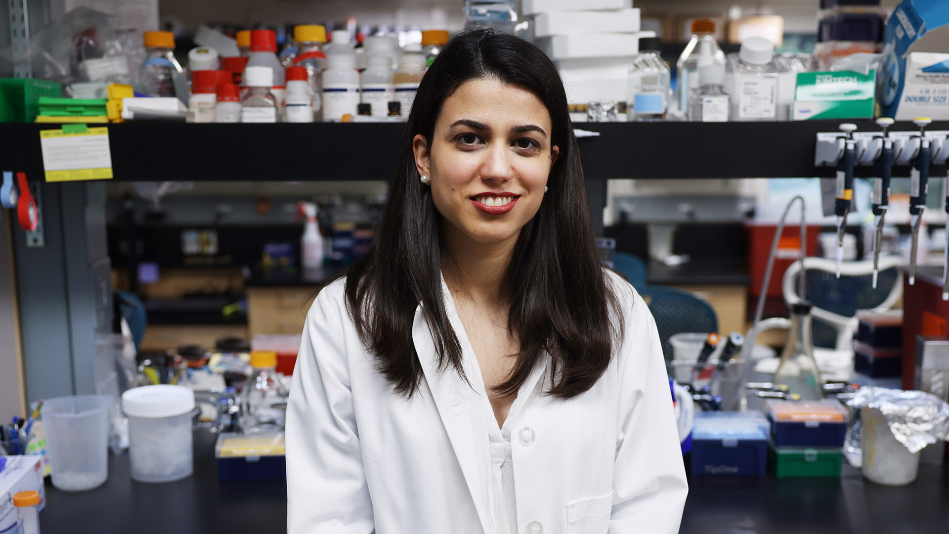Corina Amor Vegas in a white lab coat stands smiling in a laboratory filled with equipment and supplies.