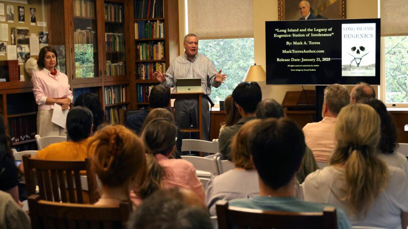 Marc Torres speaks to an audience in a library; a screen displays a book titled Long Island Eugenics.