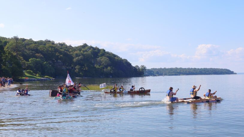 People race homemade rafts on a harbor near a wooded shoreline under a clear blue sky.