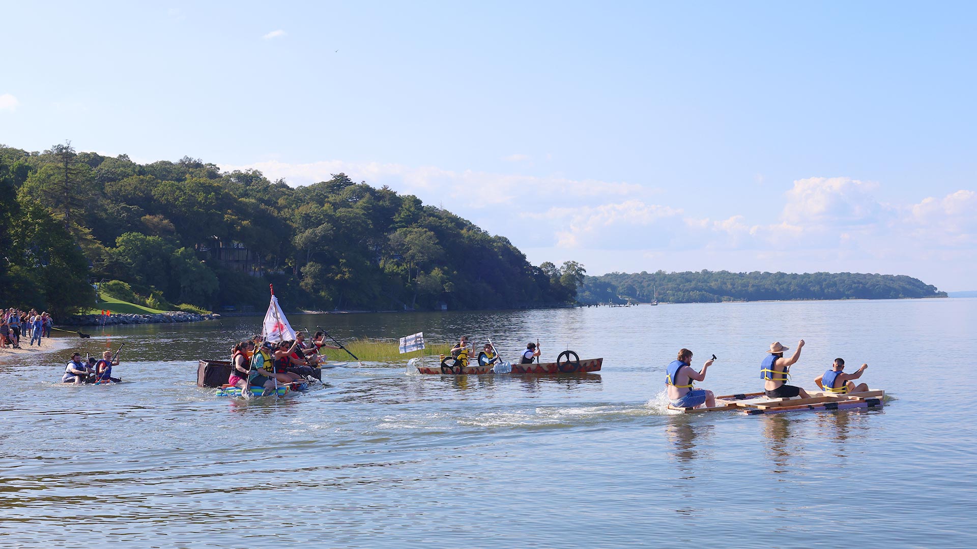 People race homemade rafts on a harbor near a wooded shoreline under a clear blue sky.