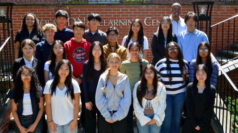 A diverse group of students and a teacher posing together outdoors on steps in front of a brick building.
