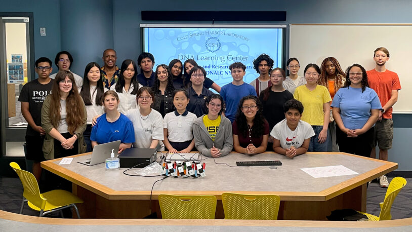 A diverse group of students and adults pose together in a classroom with a DNA Learning Center presentation screen.
