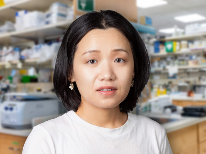 Jiaqi Zhou with short black hair stands in a laboratory, wearing a white shirt and earrings.