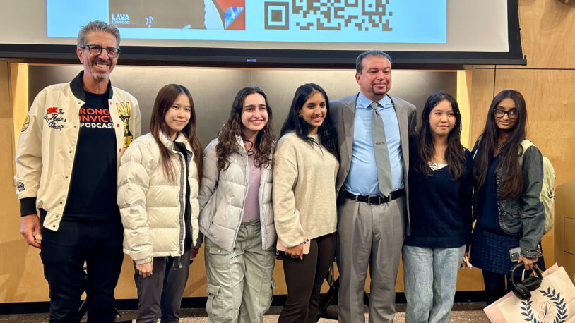 Seven people, including five young women and two men, stand smiling together in front of a screen.