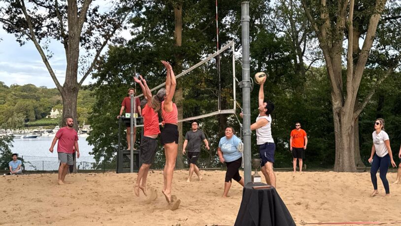 People play beach volleyball outdoors; one player jumps to spike the ball as others attempt to block.