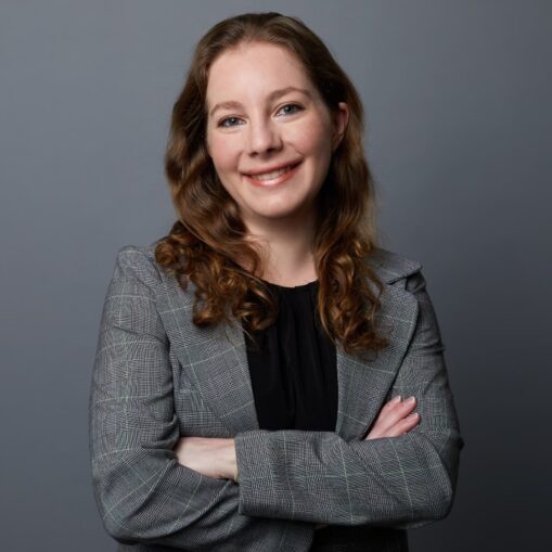 Woman with wavy brown hair in a grey plaid blazer and black top, smiling with arms crossed against a gray background.