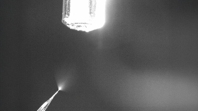 A bright nozzle sprays a fine mist or liquid downward in a dim, monochrome setting.