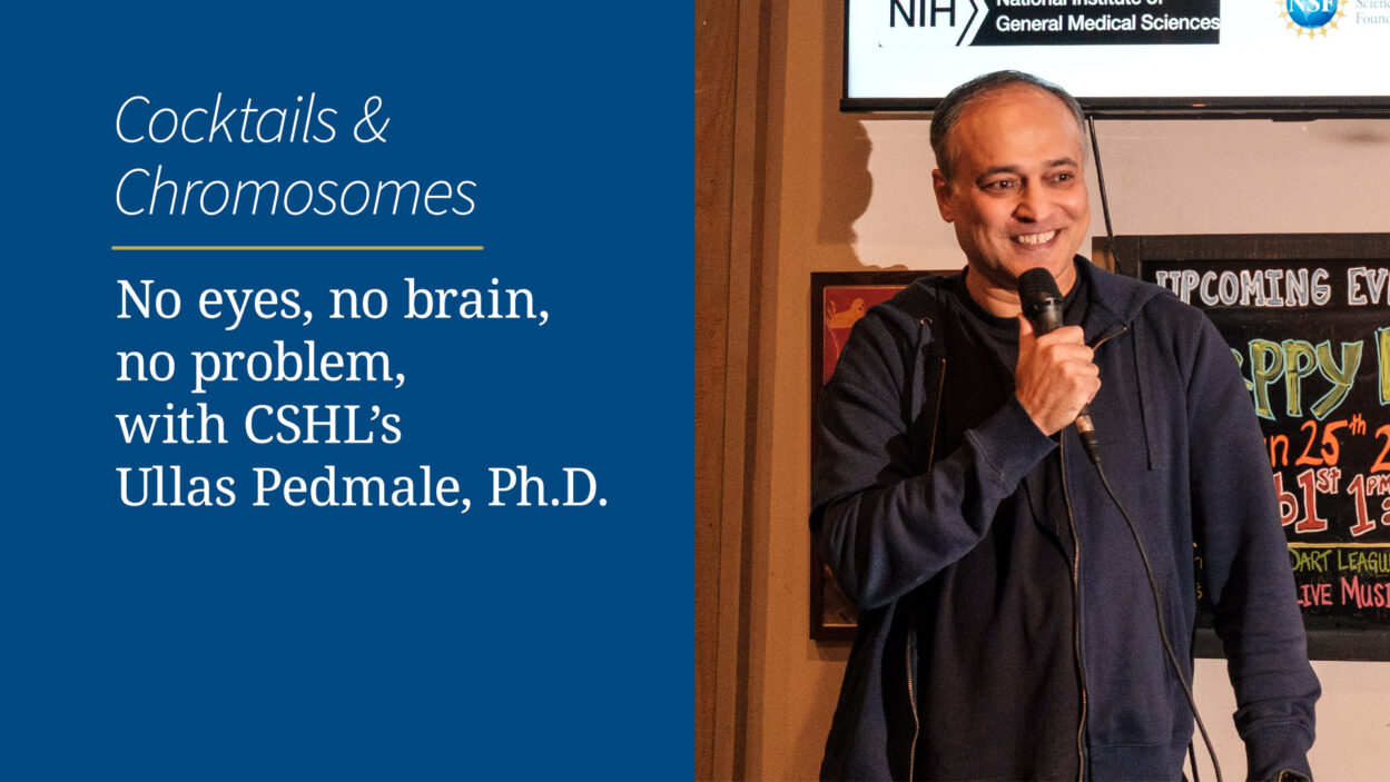 A man holding a microphone speaks next to a blue sign about a science talk featuring Ullas Pedmale, Ph.D.